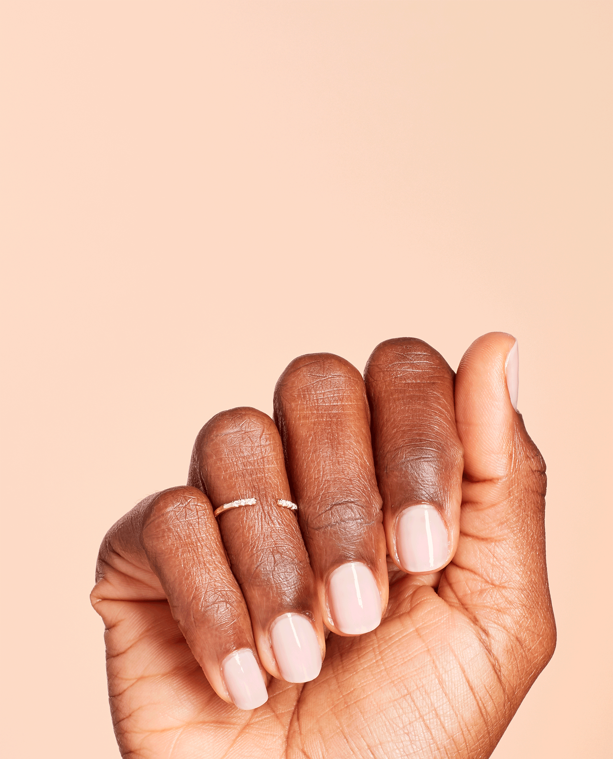 Hand with short, neatly manicured nails wears OPI Put it in Neutral nail lacquer, showing a soft pink nude shine against a peach background, ideal for promoting an elegant everyday OPI nail polish gift set in the US.